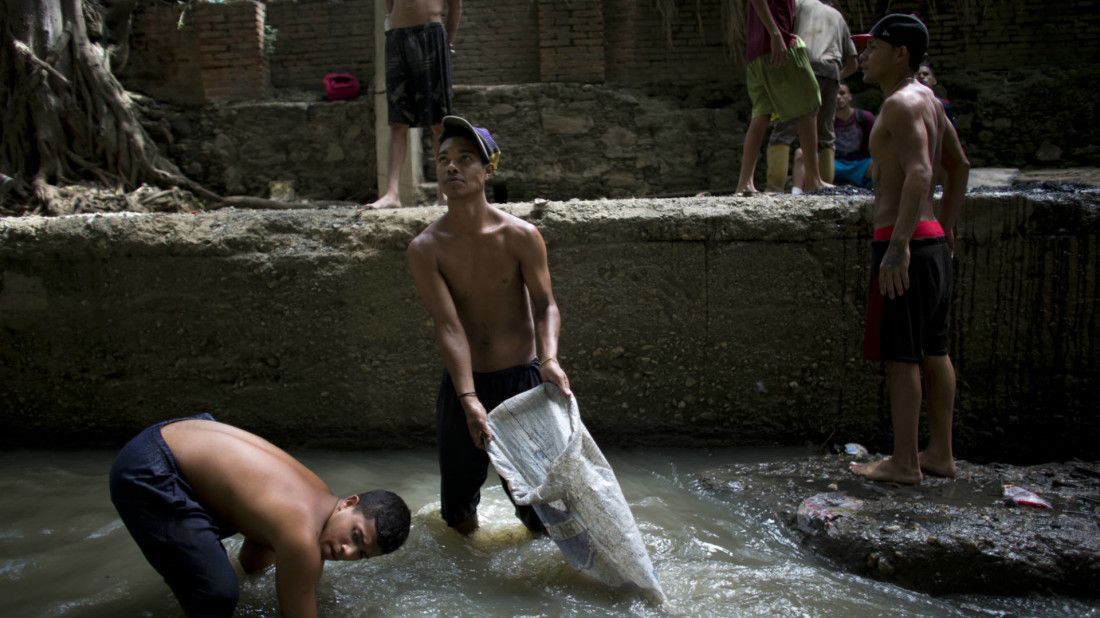 n this Nov. 30, 2017 photo, Douglas, center, holds a sack in the polluted Guaire River as he and others pull mud up from the bed of the river in search of gold and other valuables to sell, in Caracas, Venezuela. The river and the scavengers in it go largely unseen by Caracas residents speeding overhead on the city's main highway, blocked from view by concrete barriers. (AP Photo/Ariana Cubillos)